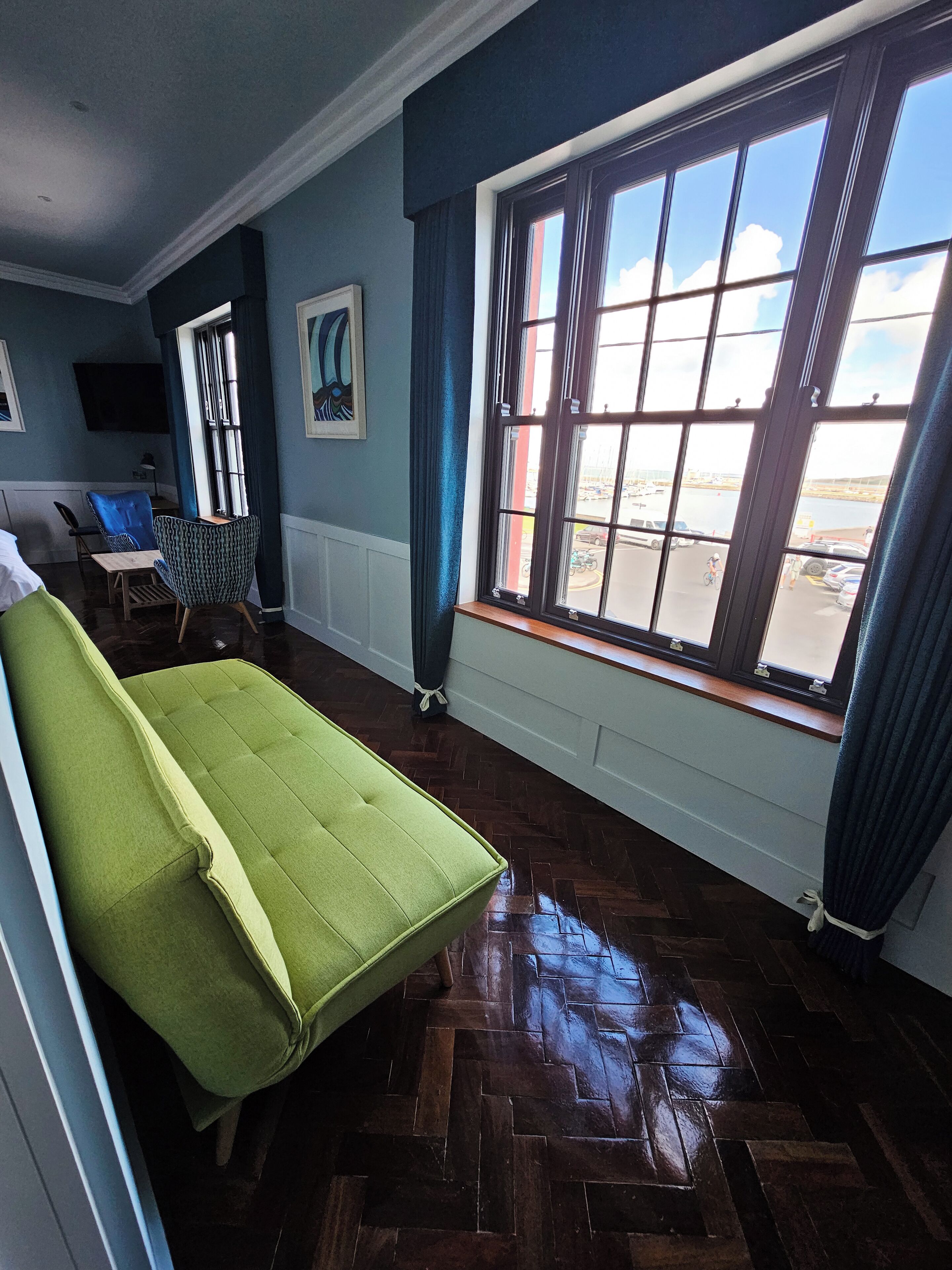 A room with a green sofa, a wooden floor, a window with a view of a marina, and a painting on the wall.