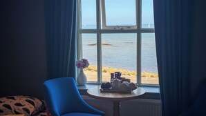 Une chambre avec un lit, une chaise, une table avec un service à thé, un vase de fleurs et une vue sur la mer par la fenêtre.