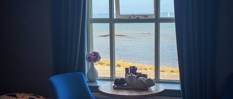 Una habitación con cama, una silla, una mesa con juego de té, un florero con flores y vista al mar a través de una ventana.