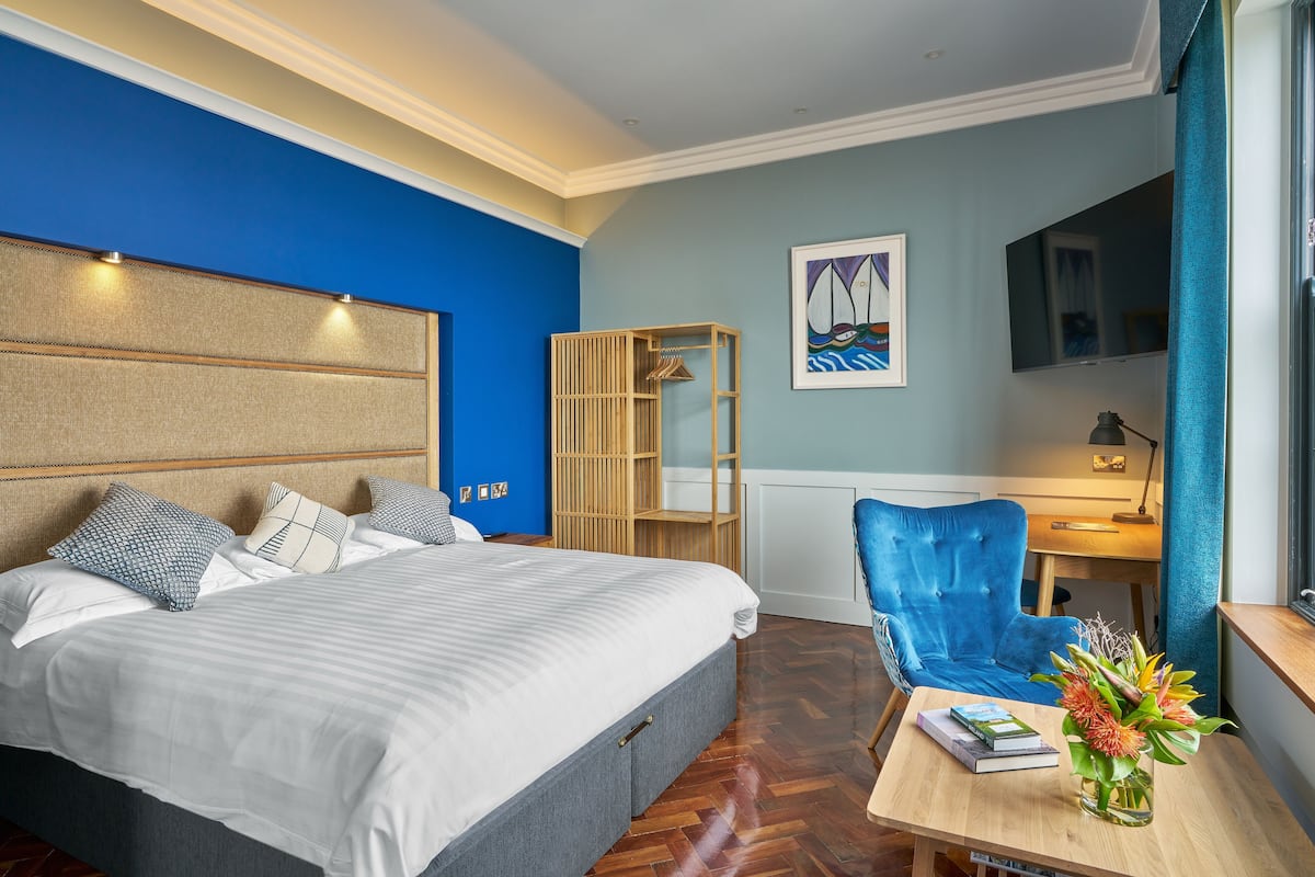 A hotel room with a large bed, a desk with a chair, a bookshelf, and a painting on the wall.