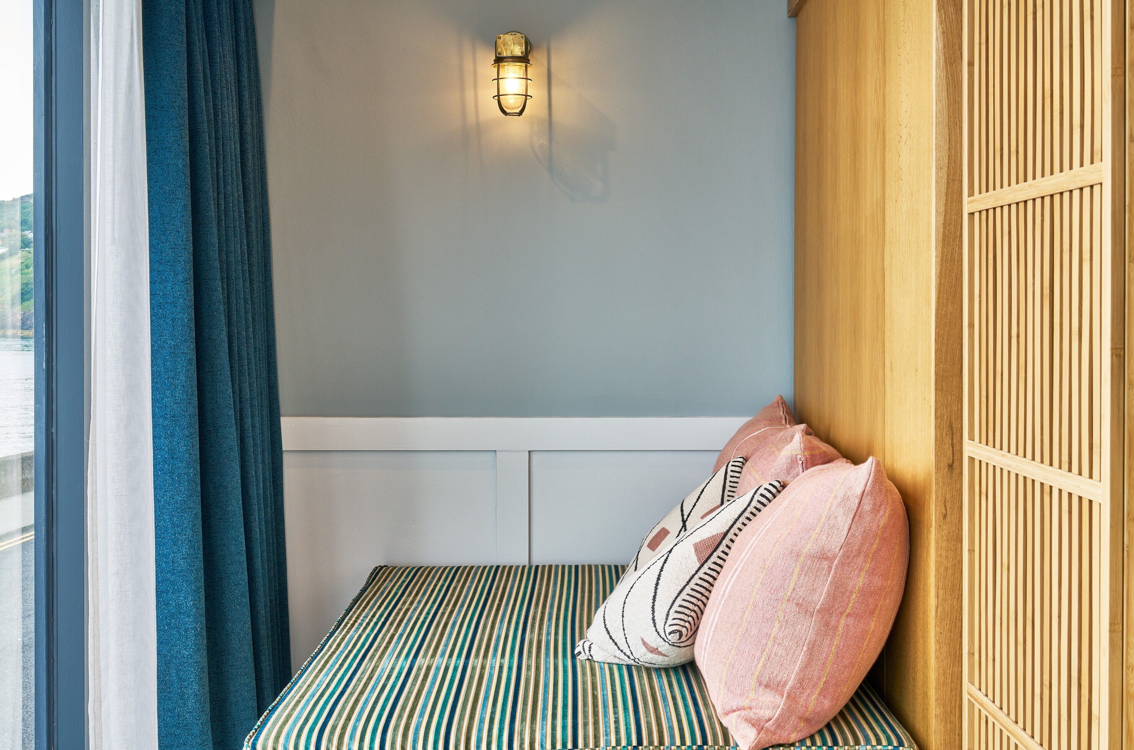 Une pièce avec un lit, une fenêtre avec des rideaux, un luminaire fixé au mur et un panneau en bois sur le côté droit.