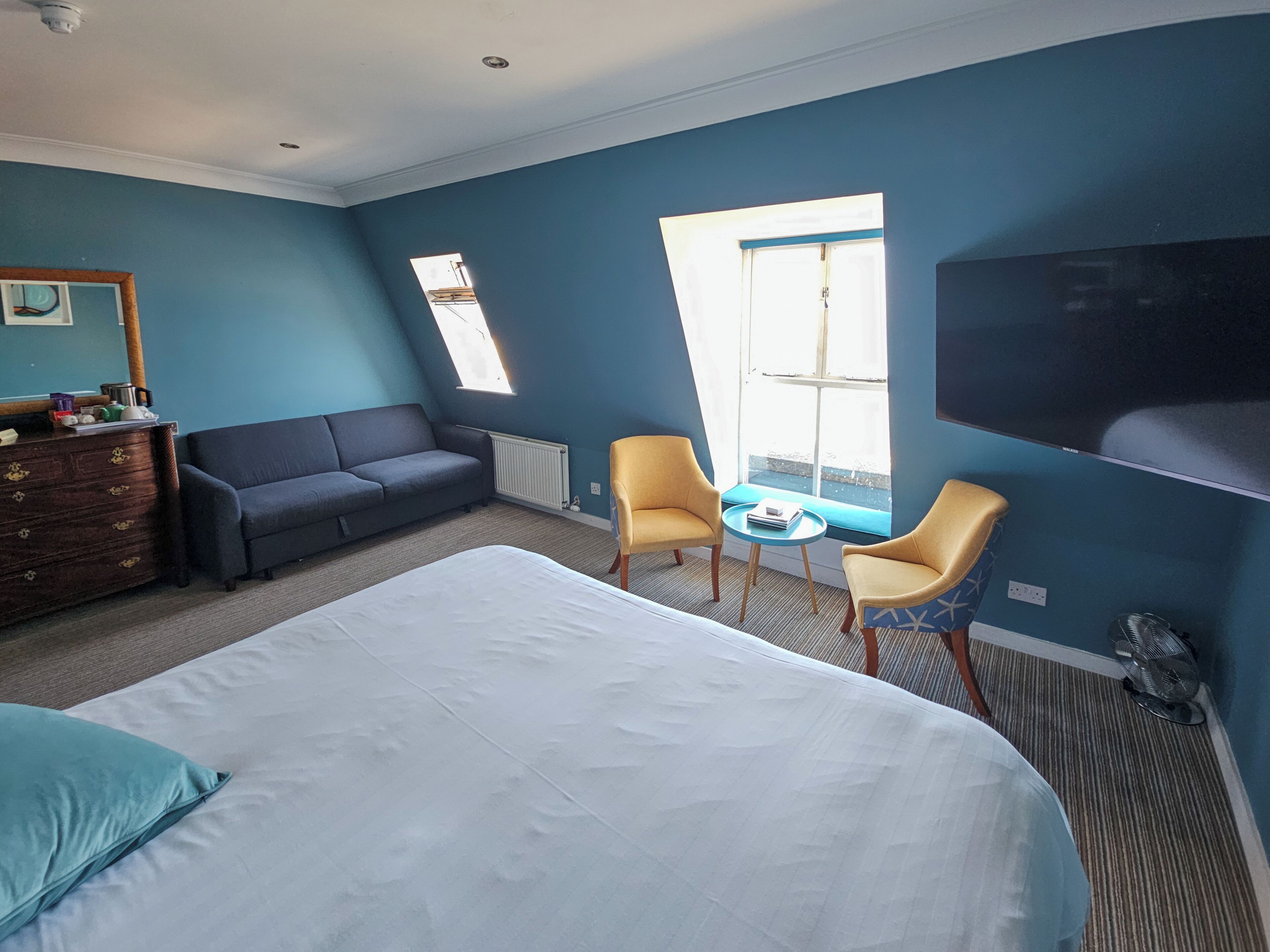A bedroom with a large bed, a dark sofa, two armchairs, a small table, a dresser, a TV, and a window with a view.
