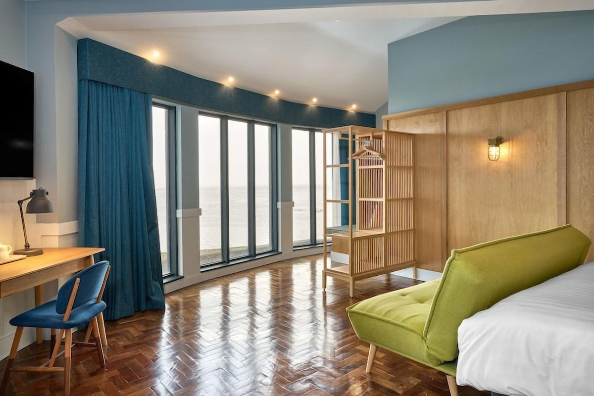 Ein Hotelzimmer mit Holzboden, einem Bett, einem Schreibtisch mit Stuhl, einem großen Fenster und Meerblick.