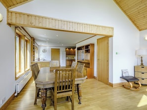 A dining area with a wooden table and chairs, a kitchen in the background, and a wooden ceiling.