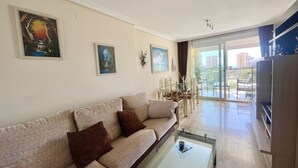 A living room with a sofa, coffee table, and dining area. There are paintings on the walls and a view of the outside through large windows. - MIRAGOLF PONIENTE DREAM R118 (Benidorm)