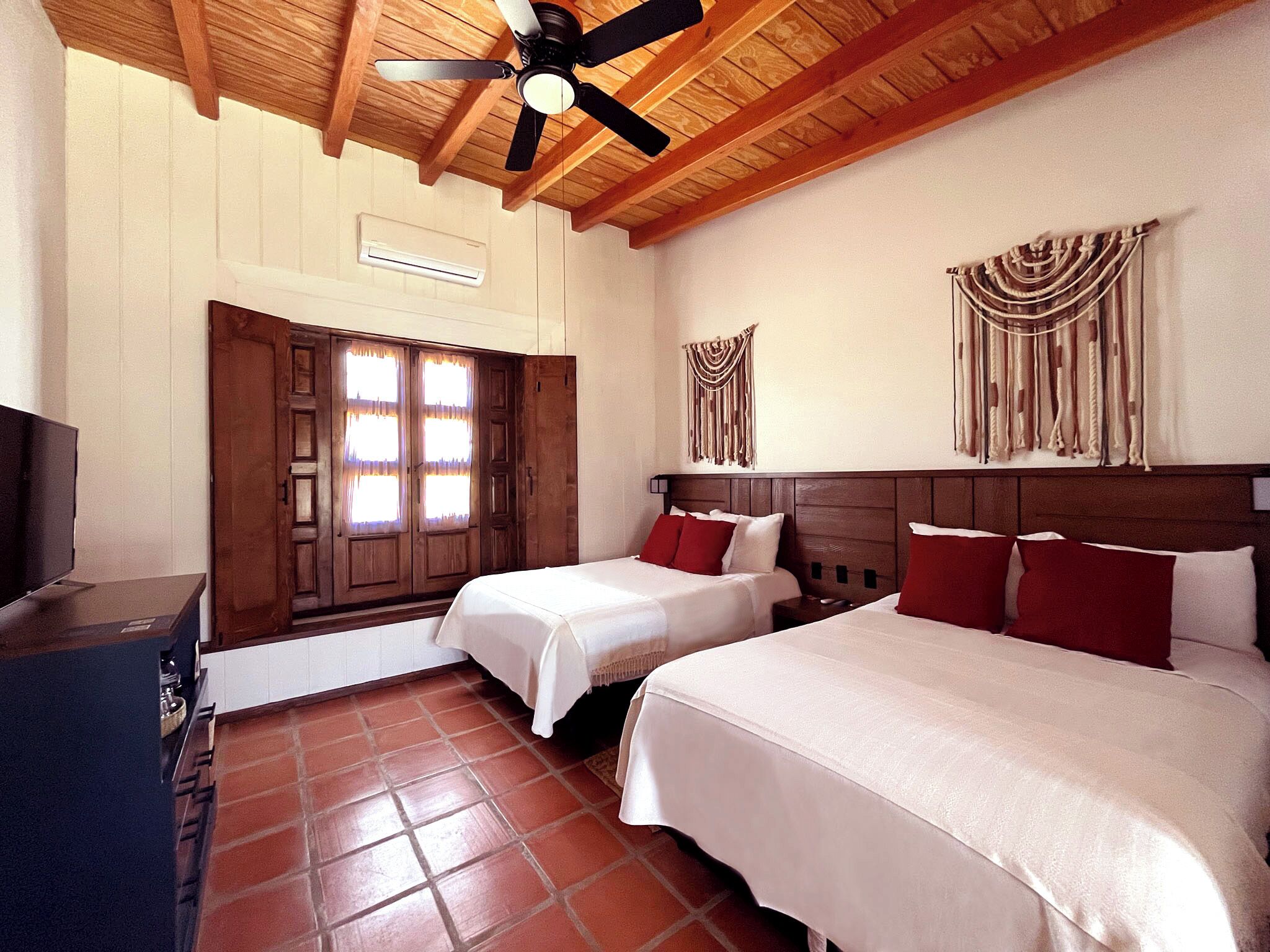 Une chambre d’hôtel avec deux lits, un ventilateur de plafond, un plafond en bois, un climatiseur fixé au mur, une porte avec des rideaux et une tapisserie murale.