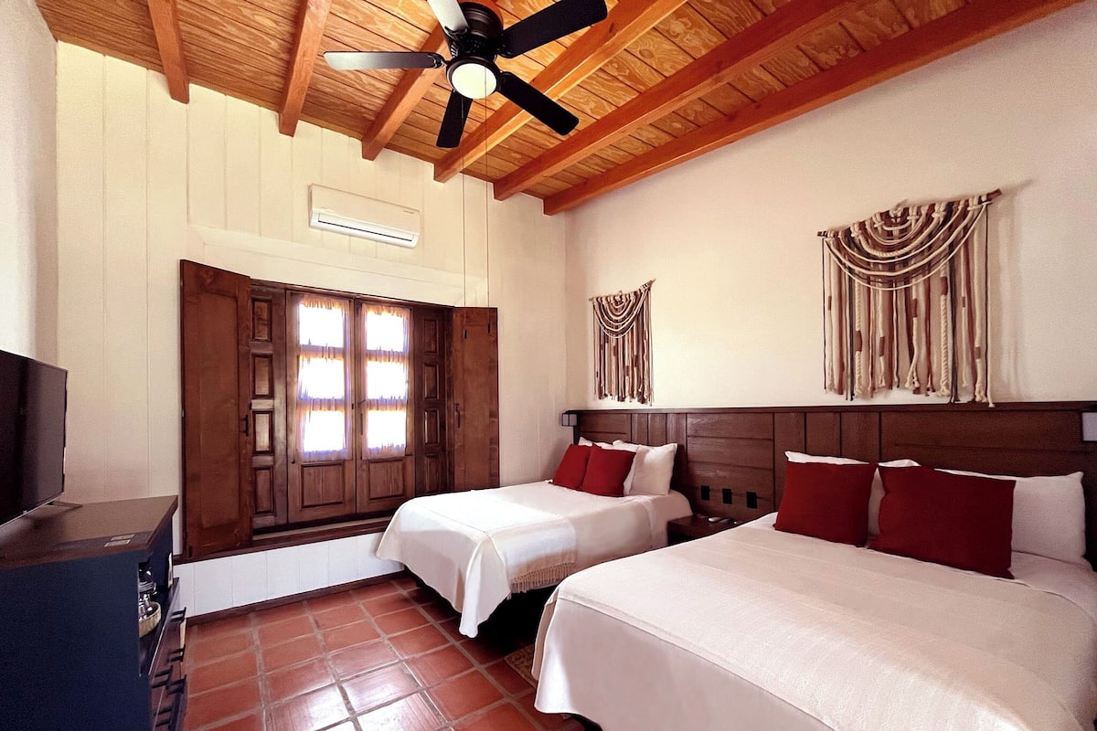 Une chambre d’hôtel avec deux lits, un ventilateur de plafond, un plafond en bois, un climatiseur fixé au mur, une porte avec des rideaux et une tapisserie murale.