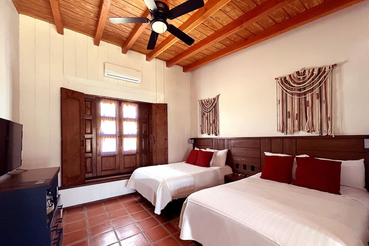 Une chambre d’hôtel avec deux lits, un ventilateur de plafond, un plafond en bois, un climatiseur fixé au mur, une porte avec des rideaux et une tapisserie murale.