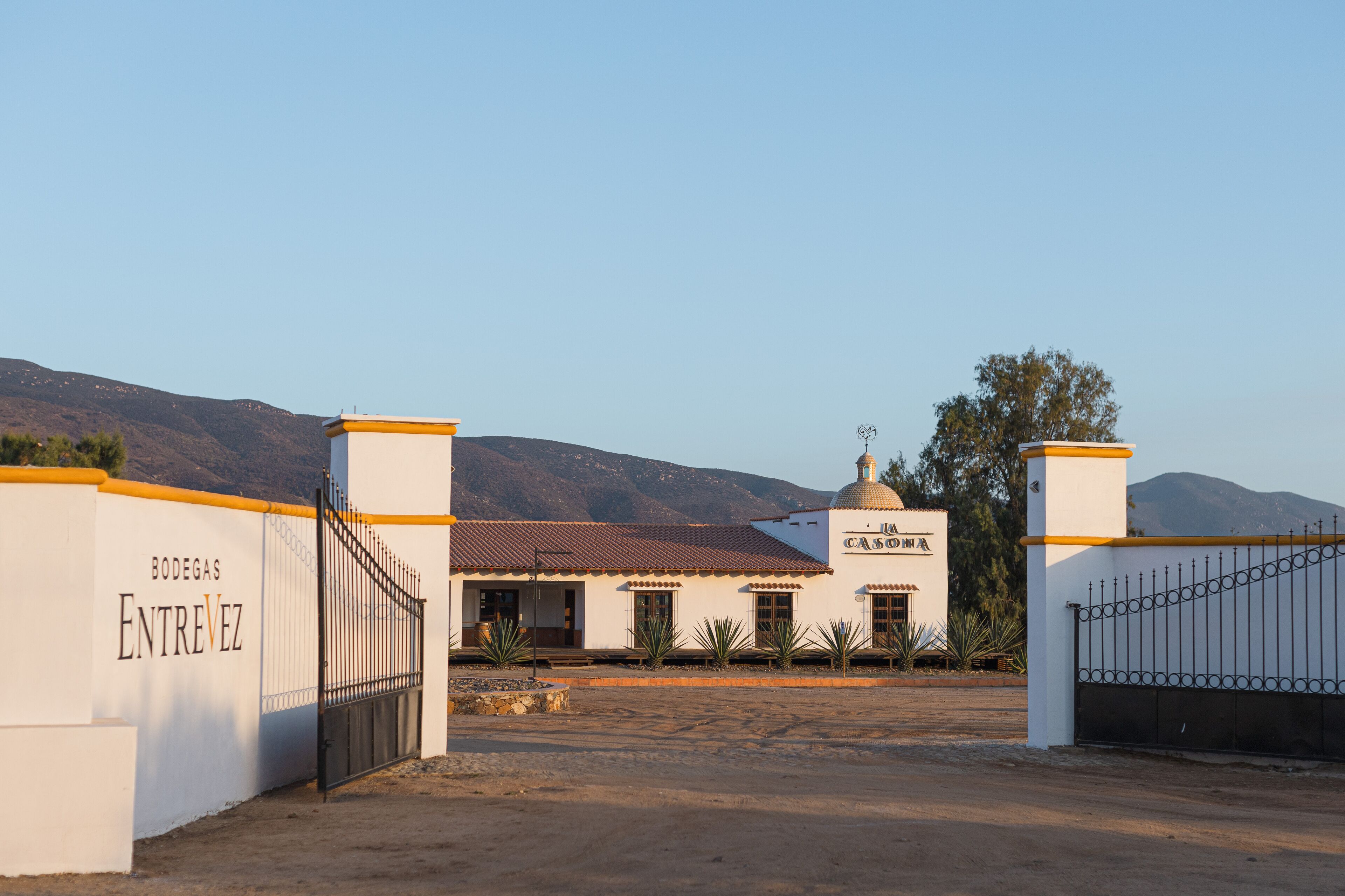 A building with a white facade and a red-tiled roof, surrounded by a fenced area with a gate labeled "BODEGAS ENTREVIZ".