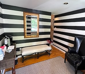 A room with striped walls, a bench, a chair, a table with items, and a window with blinds. - James Pharmacy Bed & Breakfast (Old Saybrook)