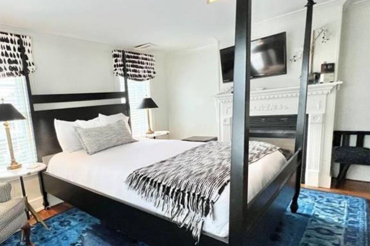 Un lit à baldaquin avec une couvre-lit noire et blanche, un tapis bleu à motifs, un téléviseur fixé au mur et une petite table avec une lampe.