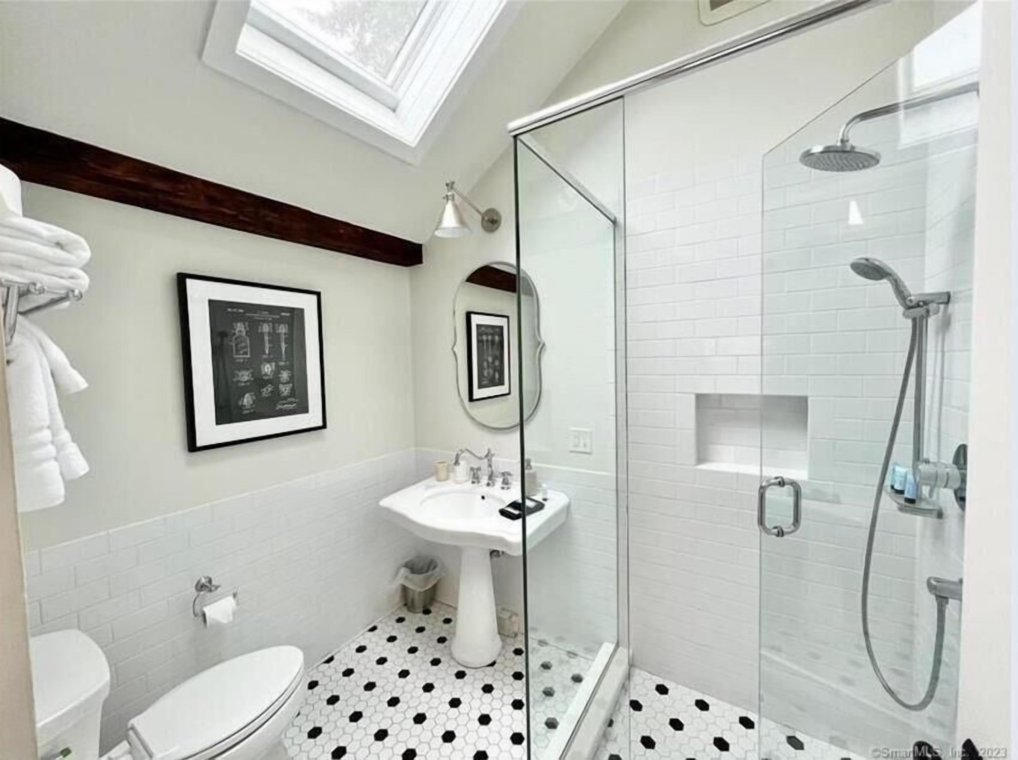 A bathroom with a white sink, toilet, and shower. There are framed pictures on the wall and a skylight above.