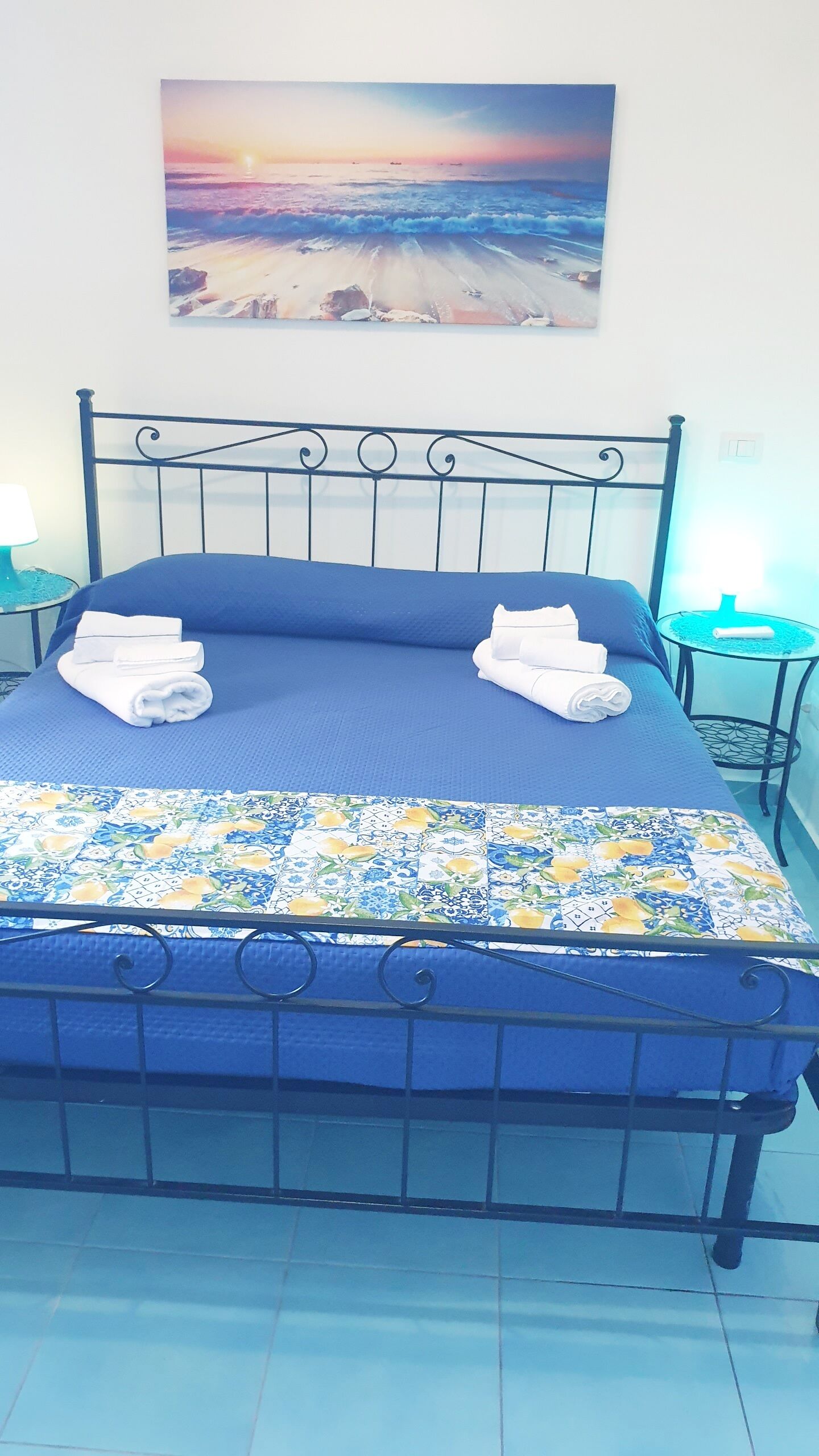 Un llit amb matalàs blau, un llençol amb dibuixos i un capçal metàl·lic, i un quadre amb un tramuntana sobre l'oceà a la paret.