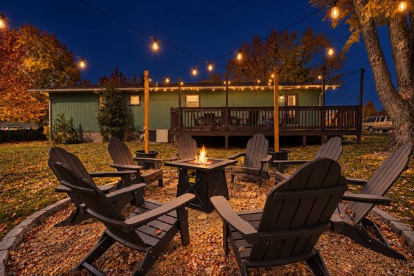 An outdoor seating area with Adirondack chairs and a fire pit, surrounded by a deck and string lights.