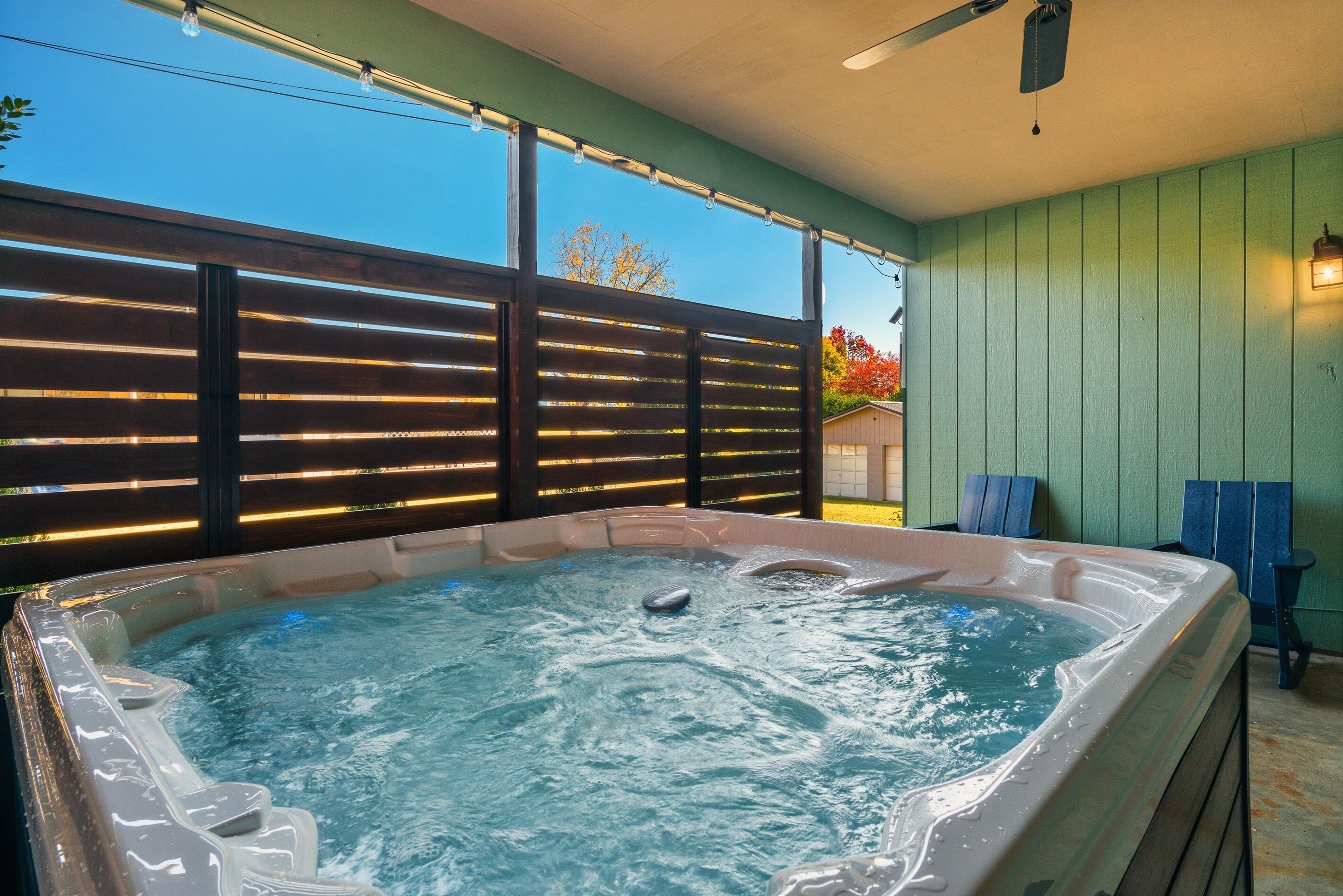 Ein Whirlpool mit Holzterrasse und Lichterkette, umgeben von einer grünen Wand und einem Fenster mit Blick auf Bäume.