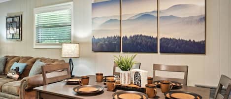 A dining area with a wooden table set for four, surrounded by chairs. There are three framed landscape paintings above the table, a lamp on the table, and a plant in a white vase.
