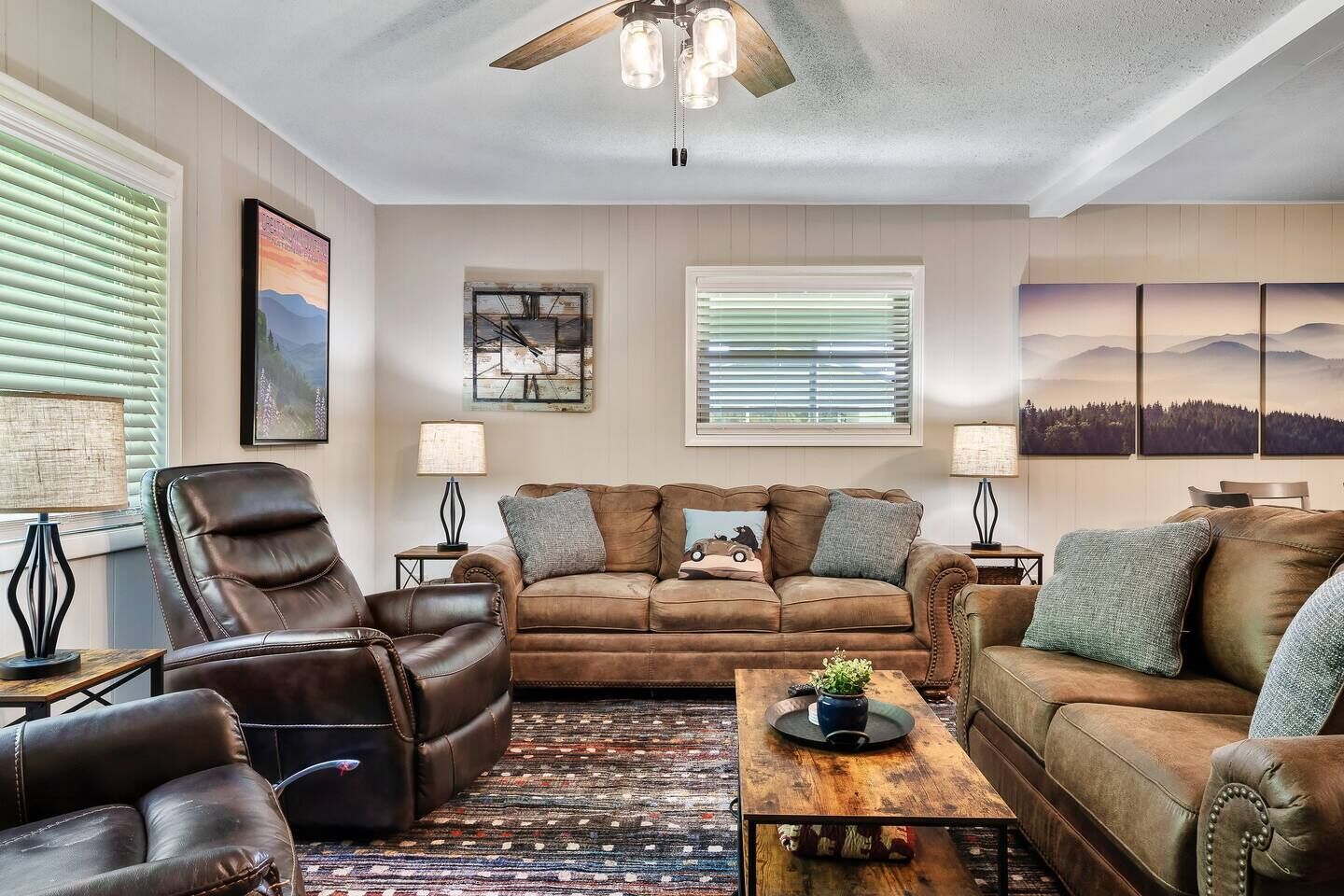 A cozy living room with a brown leather sofa, a brown leather armchair, and a wooden coffee table with a plant. There are two wall-mounted lamps and a ceiling fan.
