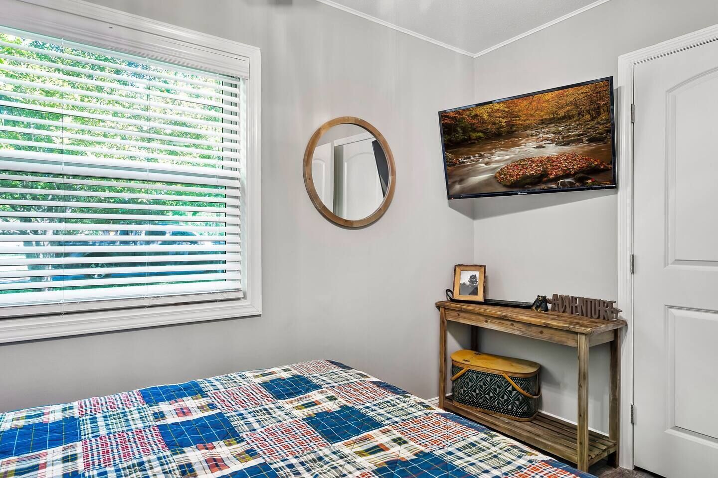 A bedroom with a bed, a window with blinds, a mirror, a wall-mounted TV, and a small wooden console table with a picture frame and a sign.