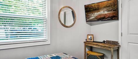 A bedroom with a bed, a window with blinds, a mirror, a wall-mounted TV, and a small wooden console table with a picture frame and a sign.