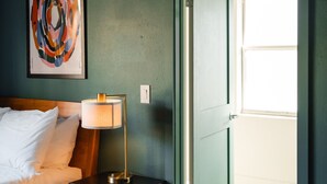 Ein Schlafzimmer mit einem Bett, einem Nachttisch mit Lampe, einem gerahmten abstrakten Gemälde und einer Tür, die auf einen Balkon führt.