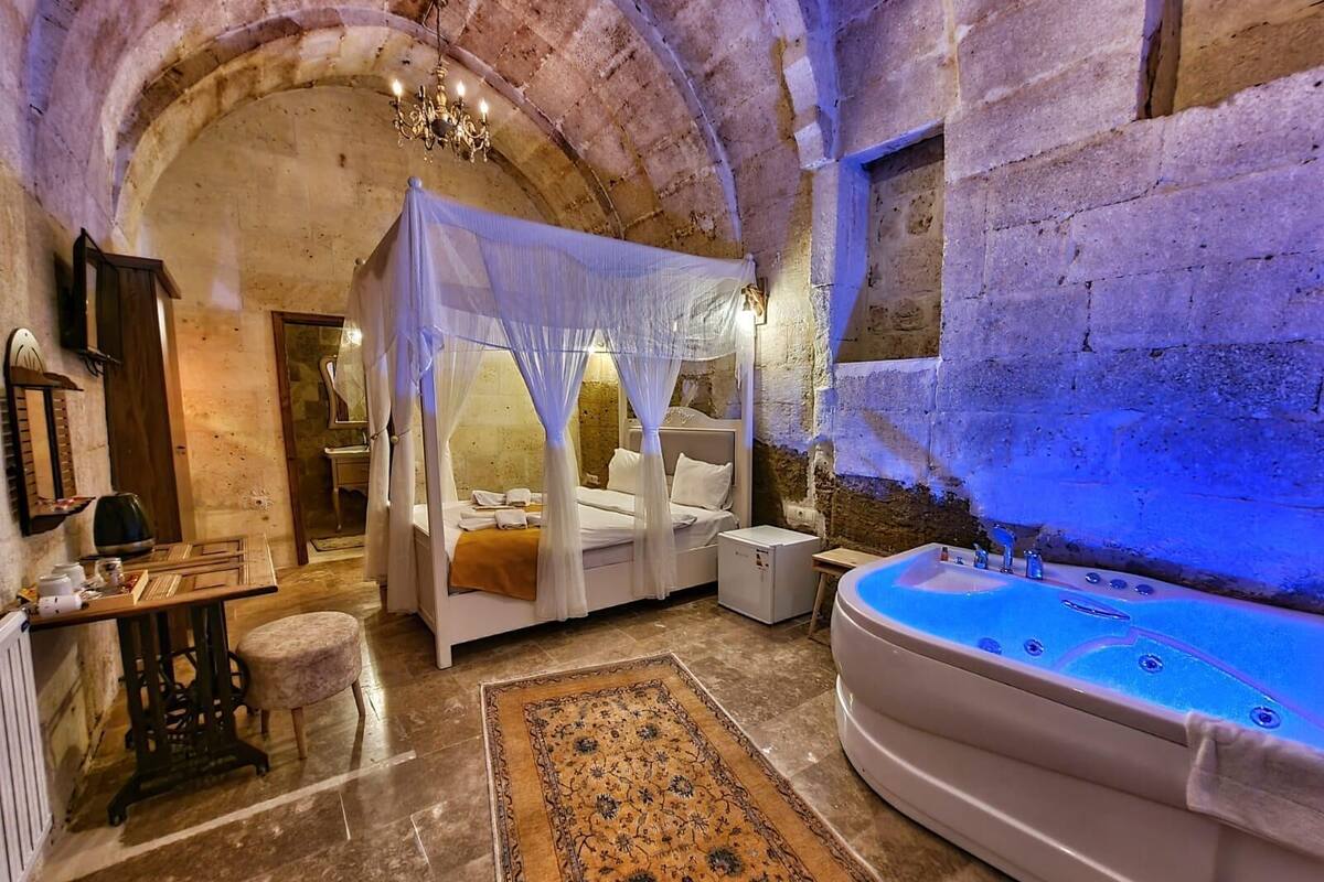 A room with a four-poster bed, a sitting area with a small table and chair, a large bathtub, and a patterned rug.