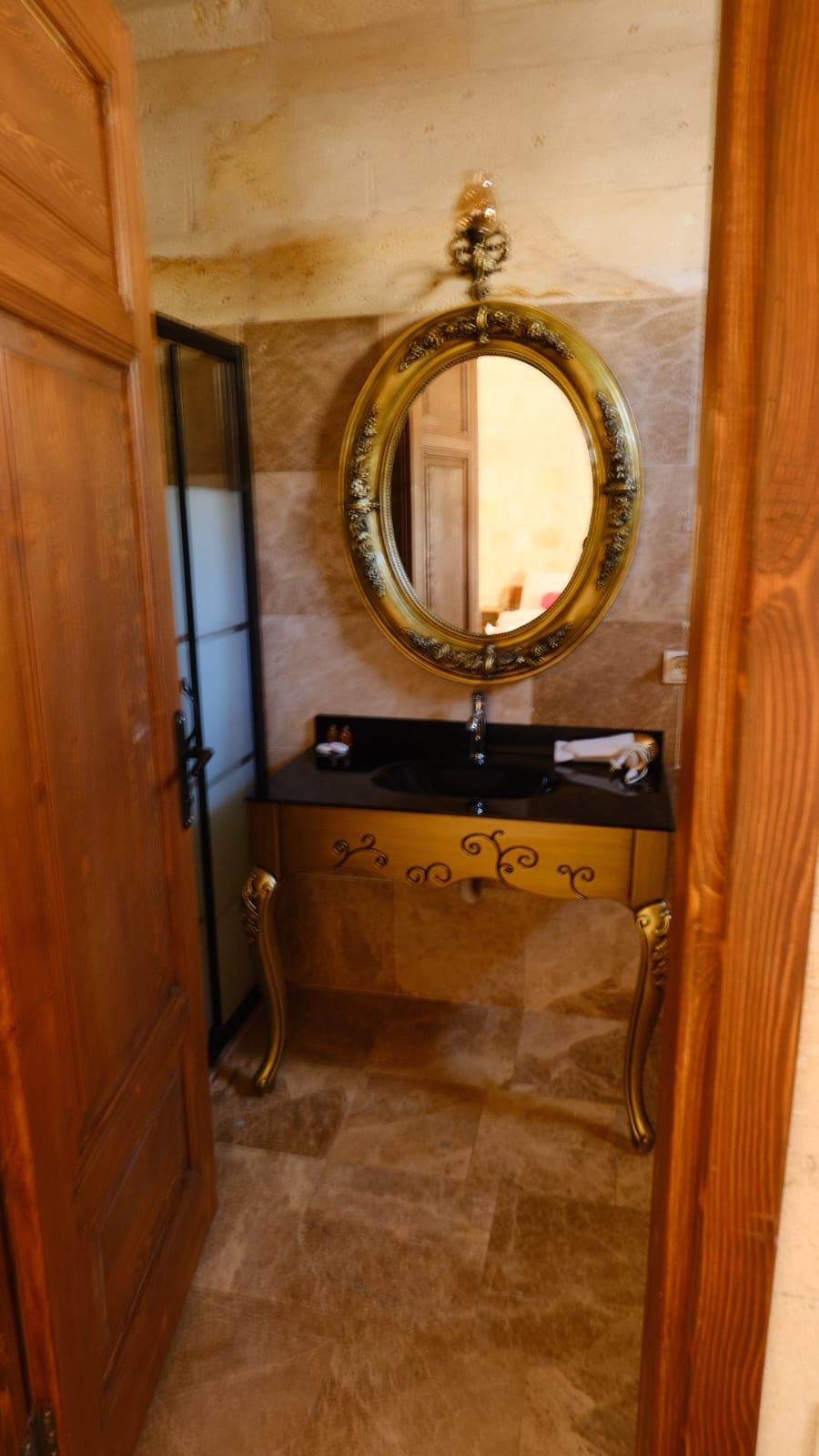A bathroom with a wooden door, a mirror with an ornate frame, a black countertop, and a decorative cabinet.