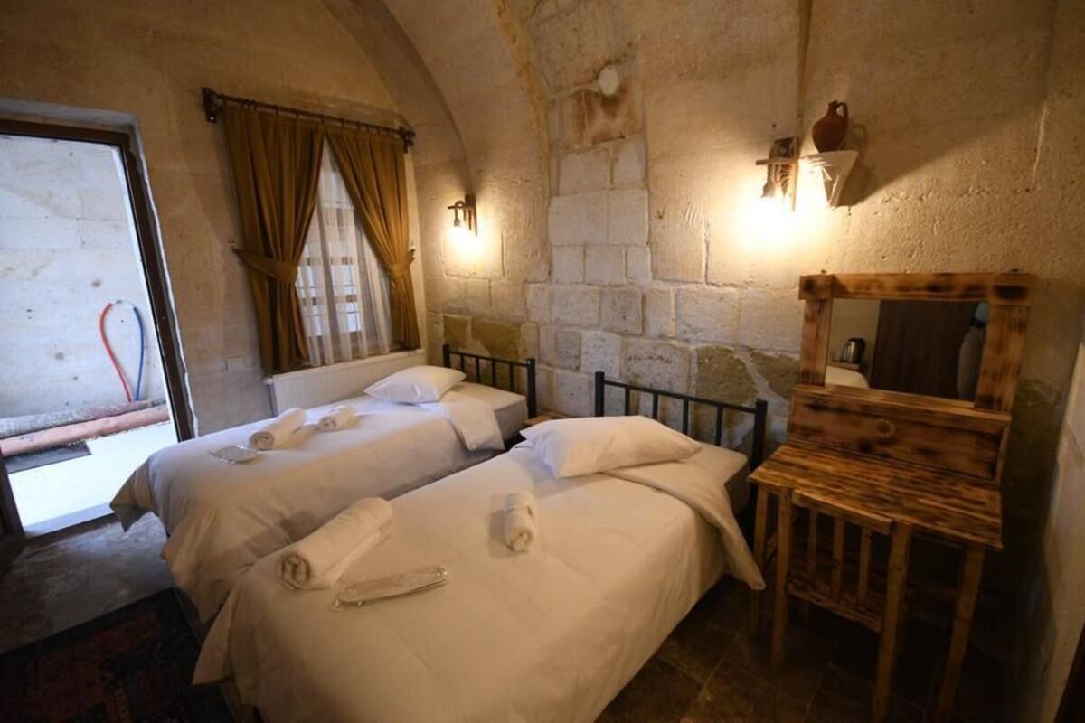 A room with two beds, a wooden nightstand, a stone wall, and a window with curtains.