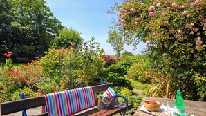 Un banc en bois surmonté d’une couverture rayée aux couleurs vives, une mangeoire à oiseaux en métal, un bol rempli de fraises et une bouteille verte, le tout posé sur une table en bois dans un jardin où les fleurs et les arbres sont en pleine floraison.