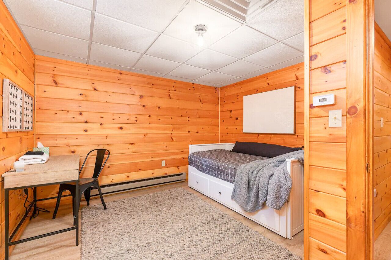 Une petite pièce chaleureuse aux murs en bois, avec un lit simple, un bureau et une chaise, et un tableau blanc accroché au mur.