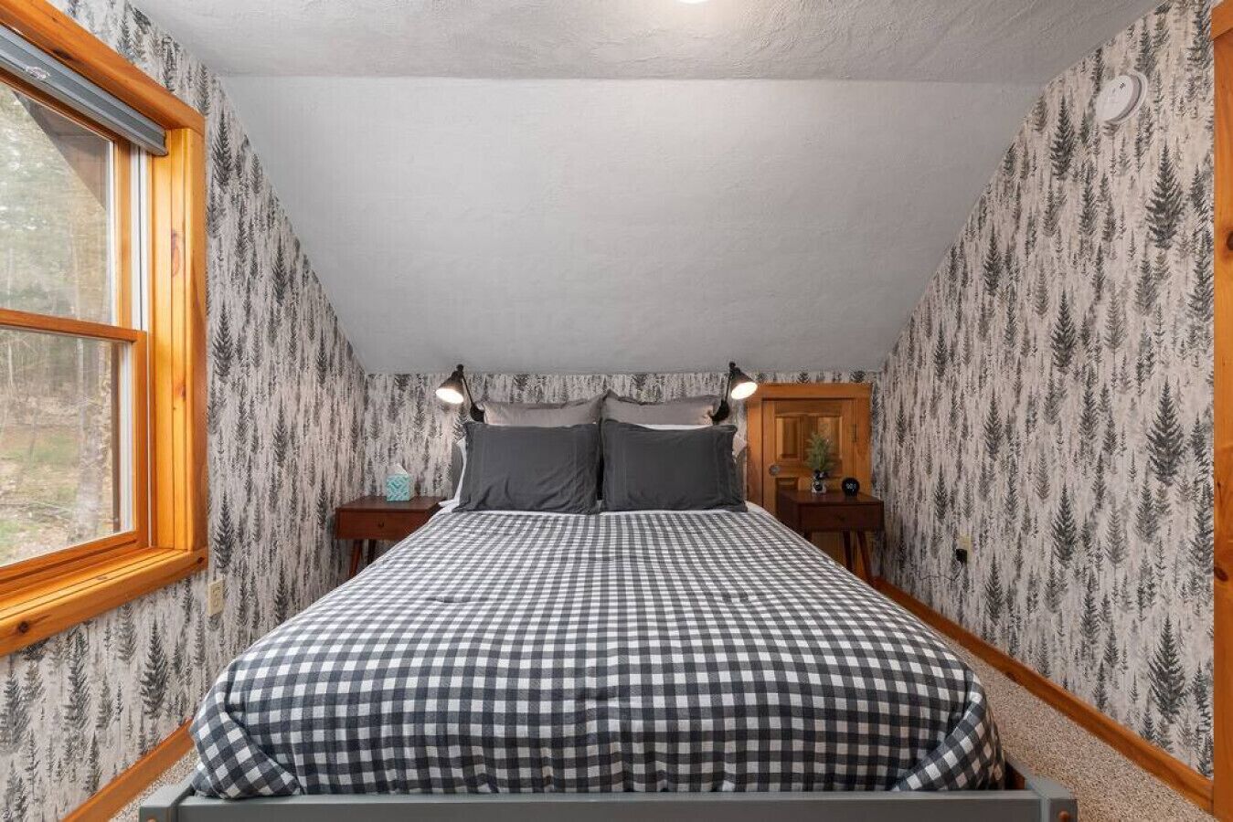 A bedroom with a checkered bedspread, two bedside lamps, a nightstand, and a window with a view of trees.