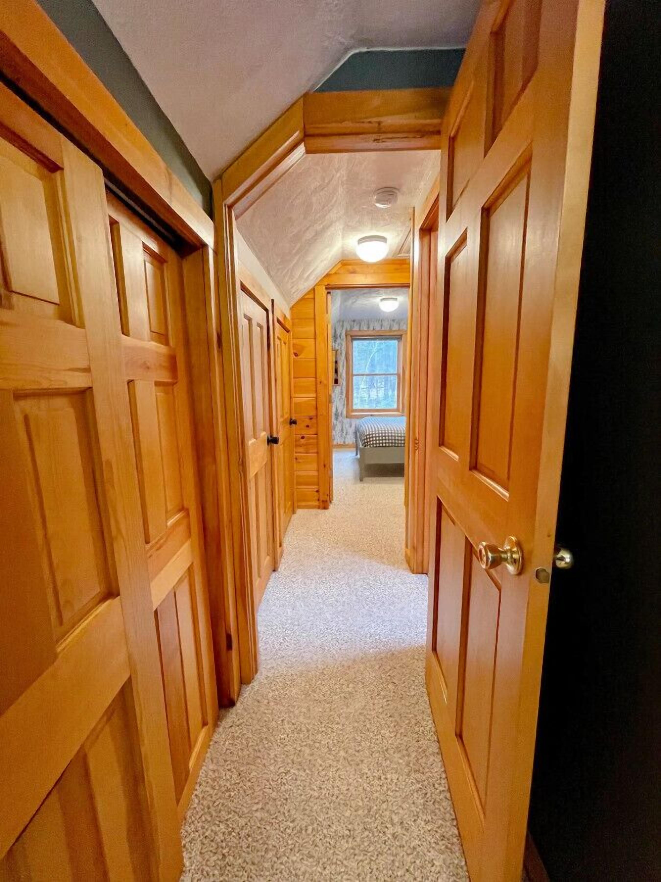A narrow hallway with wooden doors leading to a bedroom with a window and a bed.