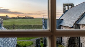 Une vue à travers la fenêtre d’un bâtiment en pierre, donnant sur un champ verdoyant, un sentier et un château situé au loin.