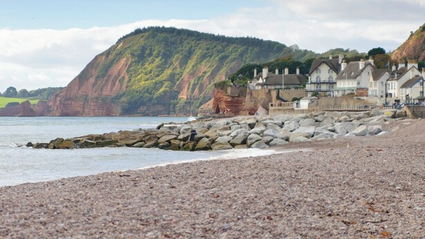 Un paisaje costero con una playa de guijarros, un rompeolas rocoso y una hilera de casas tradicionales con tejados empinados.