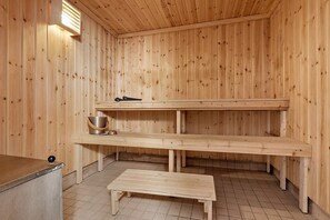 Un cuarto de sauna con paredes de madera, un banco, un estante con un balde y un par de pantuflas, y suelo de baldosas.