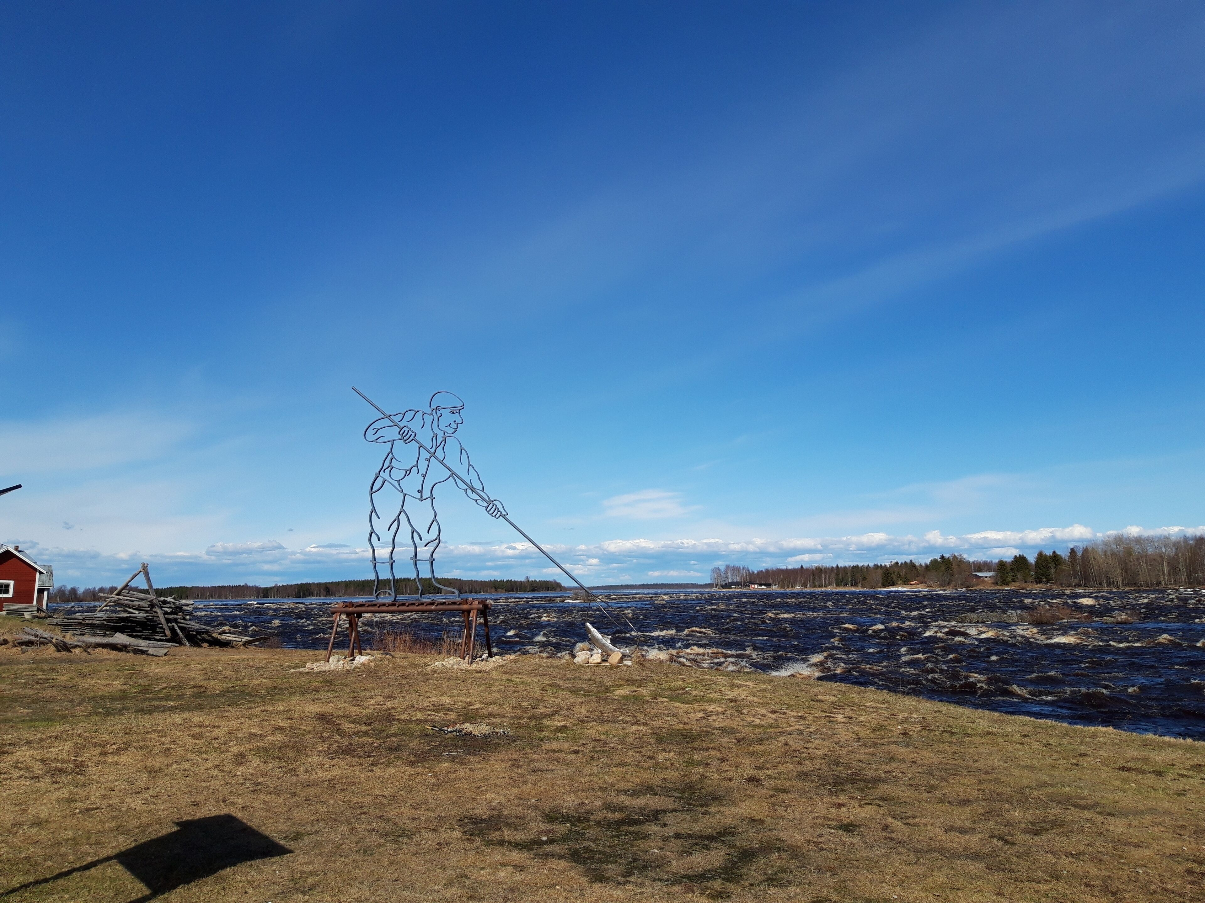 A rural landscape with a clear sky, a wooden sculpture, a red building, and a river.