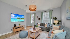 A modern living room with a grey sofa, a wooden coffee table, a blue bean bag, a TV on the wall, and a large window with curtains. - Spacious and Modern Townhome! Close to Attractions and Conventions Center! 3vc5014-10 (Orlando)