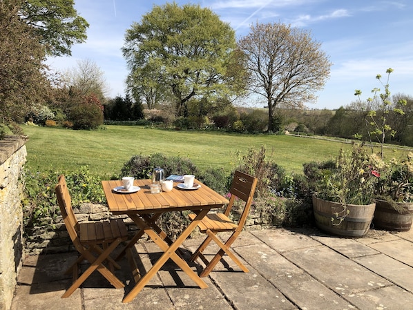 Een houten tafel met kopjes en een waterkoker, op een stenen terras met uitzicht op een gazon en bomen.