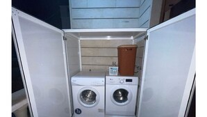 Une petite buanderie équipée d’un lave-linge et d’un sèche-linge à chargement frontal, d’une petite étagère avec un panier et d’un panneau de commande.