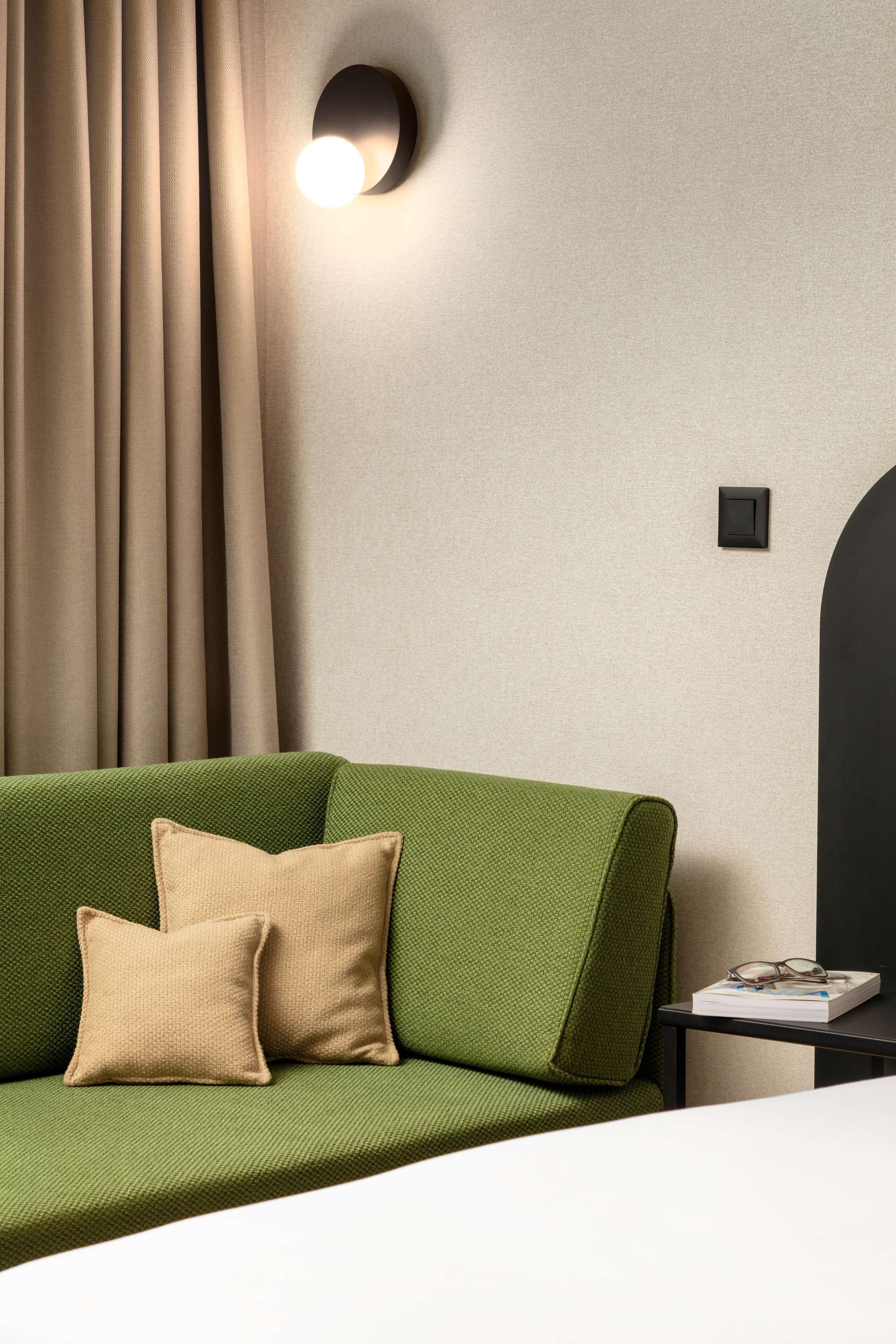A green sofa with beige cushions, a small table with a book and glasses, and a wall-mounted light fixture.