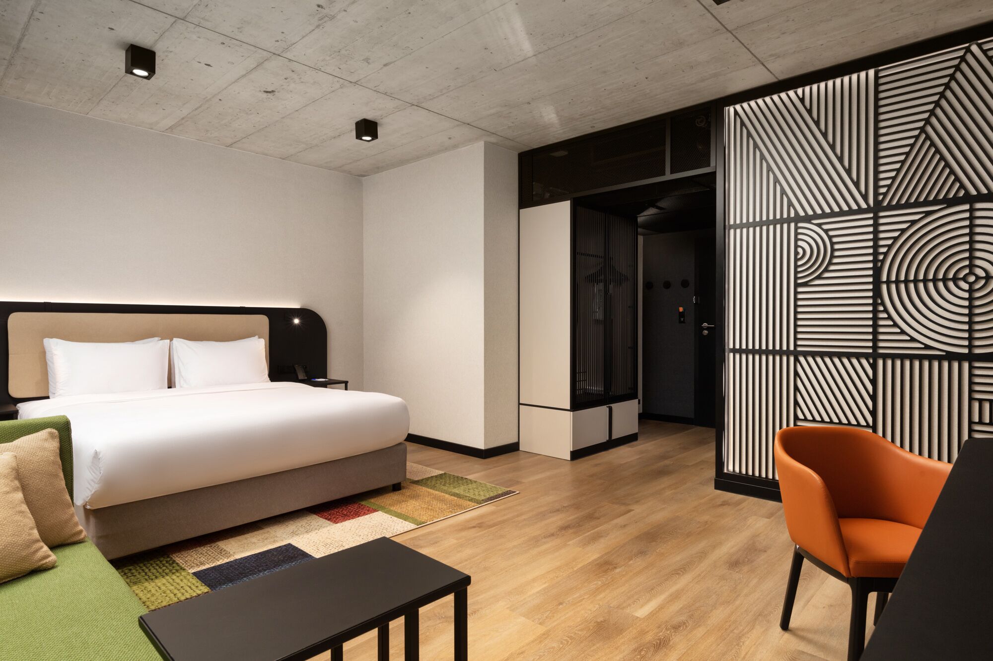 Una habitación de hotel moderna con una cama grande, un sofá verde, una mesita pequeña y un panel decorativo en la pared.