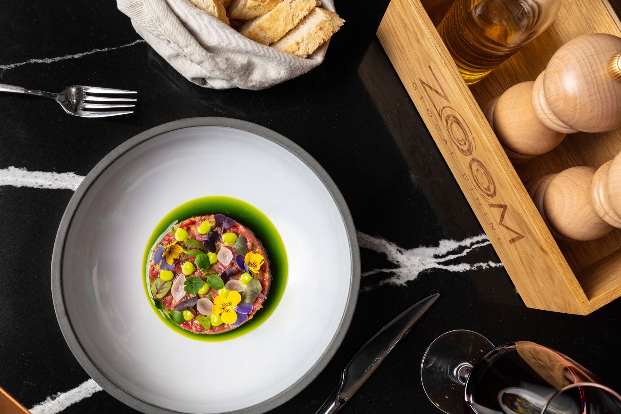 Una mesa de comedor con un plato de comida, un tenedor, un cuchillo, una copa de vino y una caja de madera con pan.