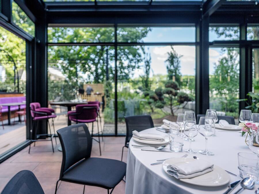 An outdoor dining area with a table set for four, surrounded by chairs, and a glass enclosure with a view of greenery.