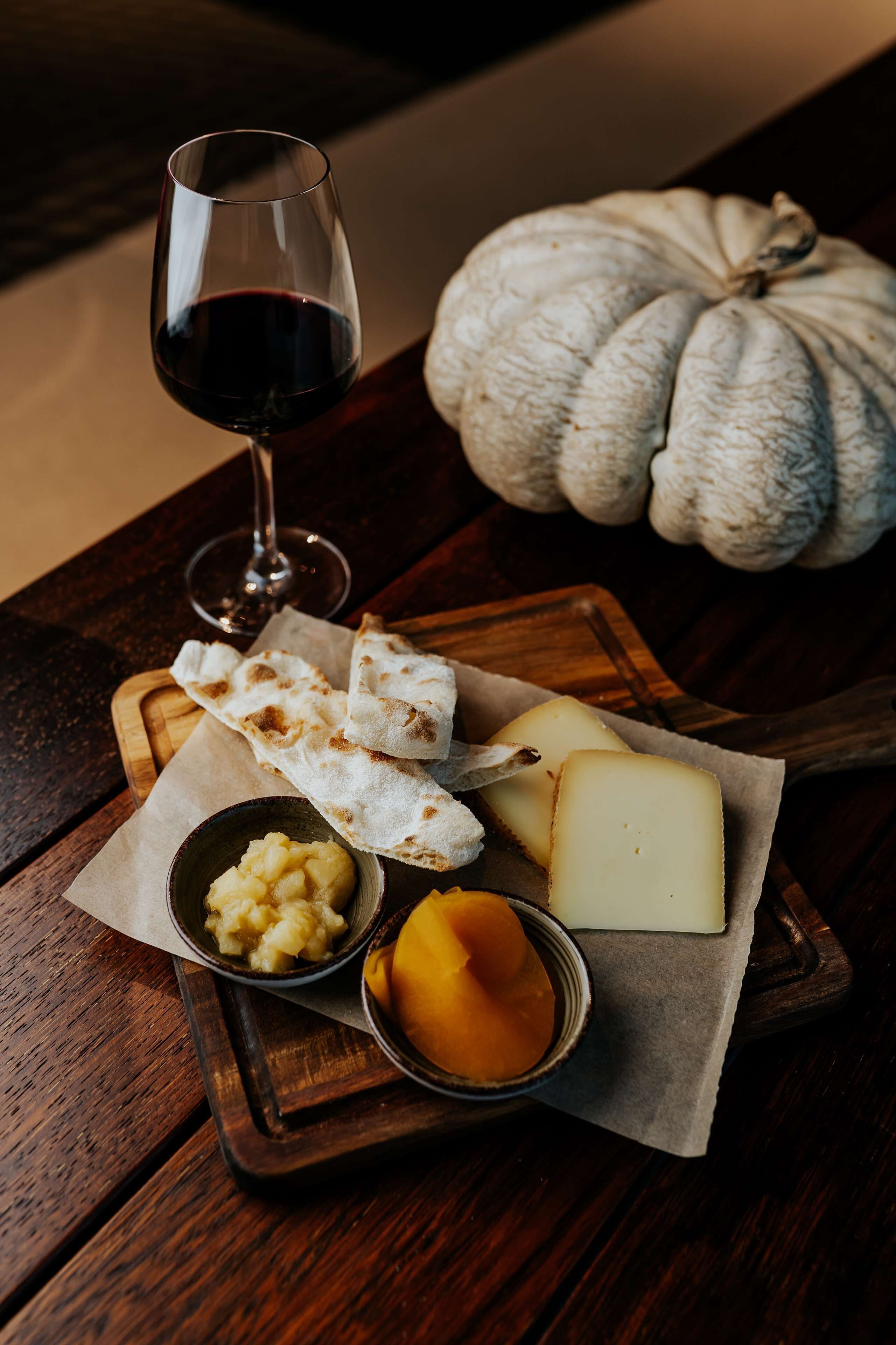 Una mesa de madera con una tabla de quesos, una copa de vino tinto y una calabaza.