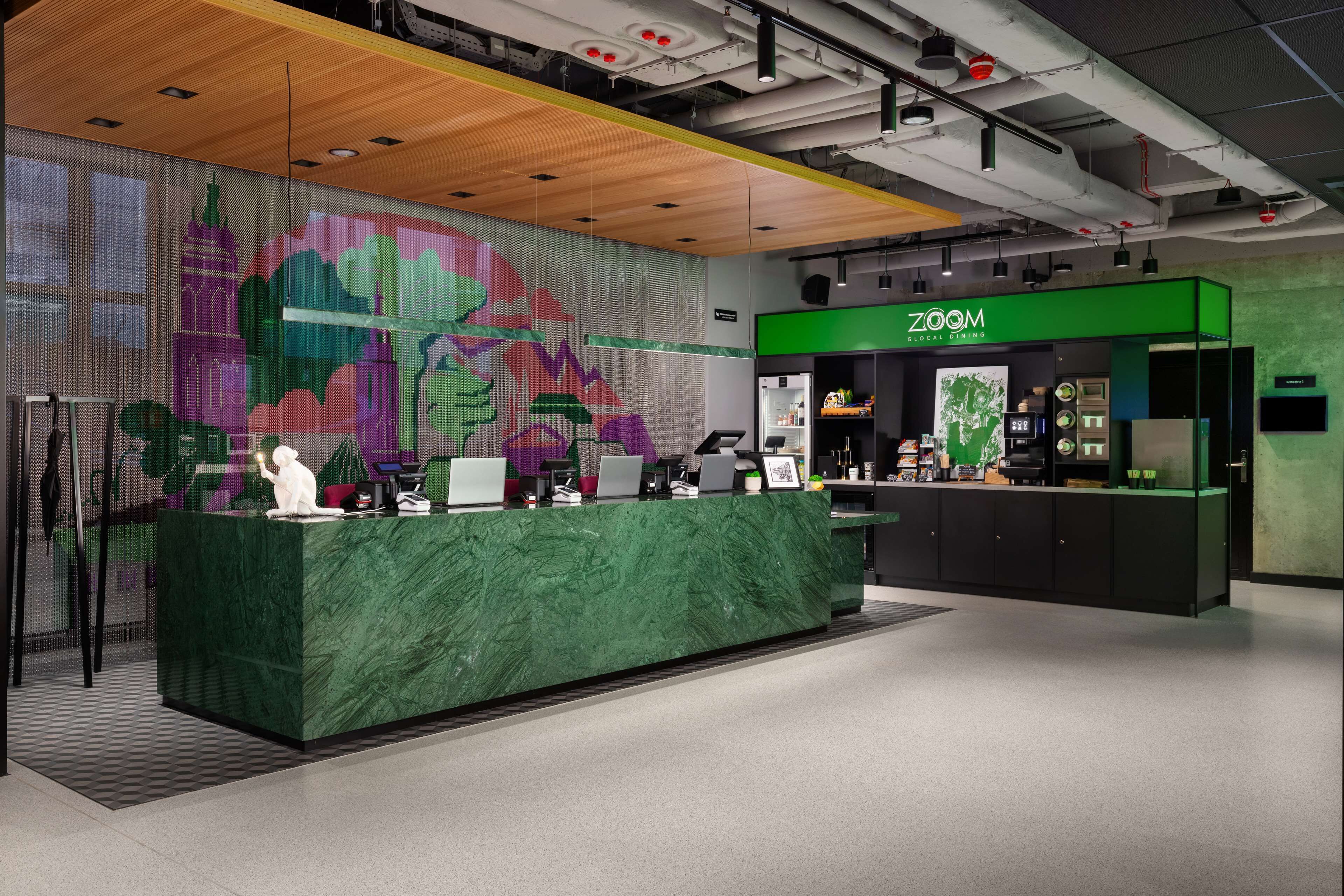A modern reception area with a green marble counter, a large digital screen displaying colorful graphics, and a green sign reading "ZOOM".