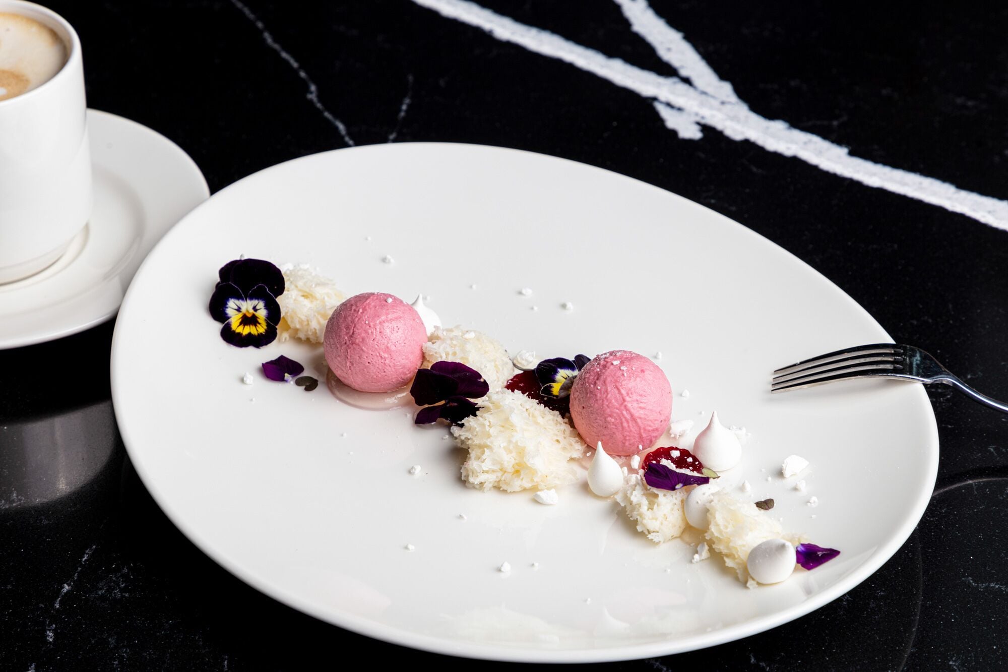 A dessert plate with pink ice cream and white coconut shavings, accompanied by a fork and a cup of coffee on a black marble table.