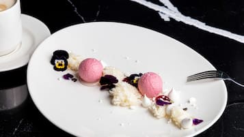 Un plato de postre con helado rosa y coco rallado blanco, acompañado por un tenedor y una taza de café sobre una mesa de mármol negro.