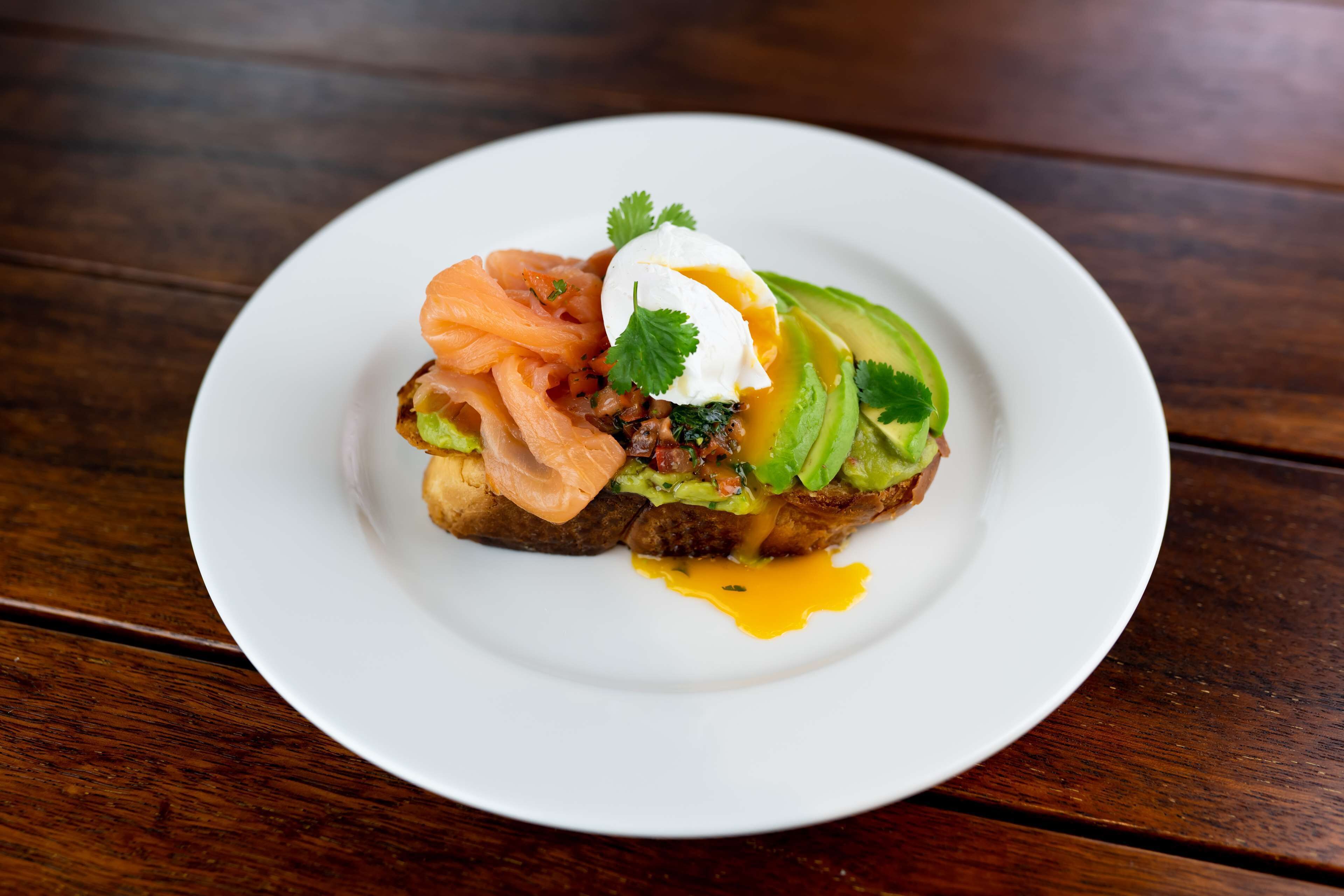 A plate with a sandwich, avocado, and a poached egg on a wooden table.