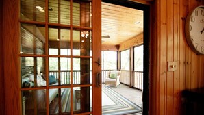 Un intérieur en bois avec une grande fenêtre, une horloge et une vue sur une véranda aménagée avec du mobilier d’extérieur.