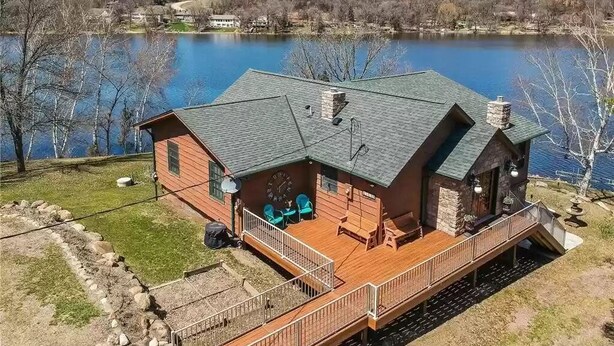 Une cabane dotée d’une terrasse donnant sur un lac, d’une cheminée en pierre et d’un petit espace porche aménagé avec des sièges d’extérieur.