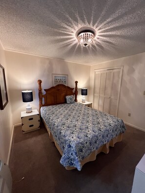 Quarto com cama de madeira, duas mesas de cabeceira com luminárias, luminária de teto, um guarda-roupa e um quadro na parede.